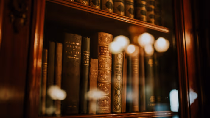 Close-up of vintage leather-bound books with ornate spines inside a wooden bookshelf with glass reflecting lights.